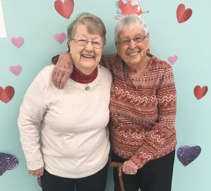 2 smiling older women standing in front of wall with hearts.