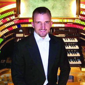 man standing in front of Organ with 4 rows of piano keys and hundreds of brightly colored switches