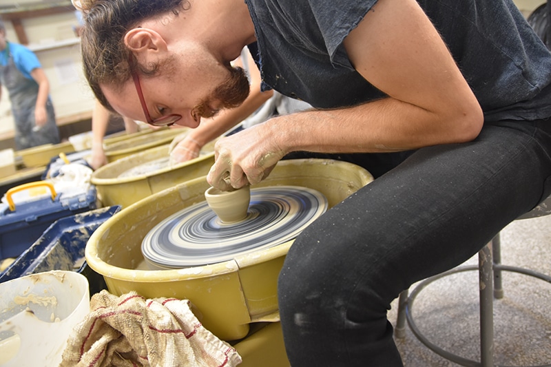 man leaning over a pottery wheel with small cup spinning and being formed with his hands.