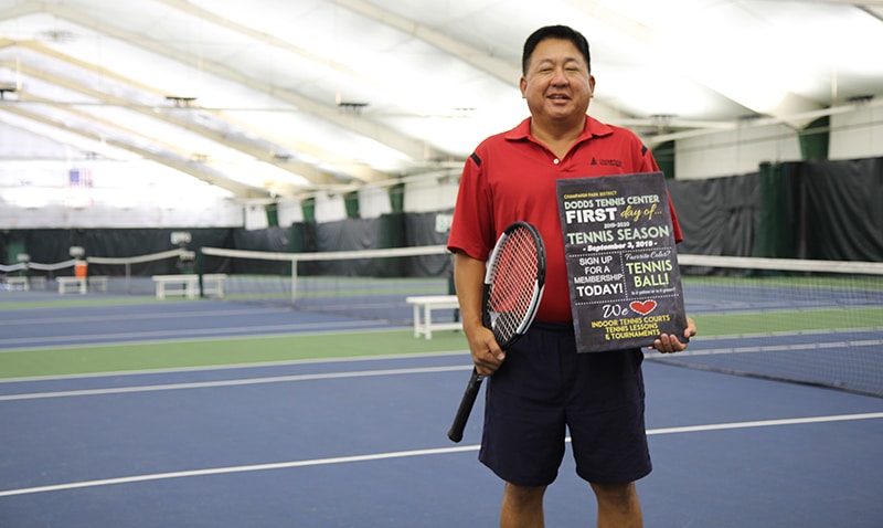 Yuri Sohn standing in front of indoor tennis courts holding a tennis racket and sign that says: Dodds Tennis Center First Day of Tennis Season.