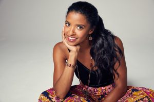 Renee Elise Goldsberry in front of grey backdrop, sitting, smiling with elbow on knee