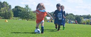 little girl in orange jersey running down the soccer field kicking a soccer ball with 2 boys in dark blue jerseys chasing her down.
