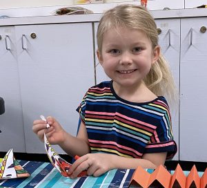 Young girl sitting at table smiling straight to the camera while coloring fish paper lantern she is making.