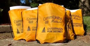 5 orange drawstring bags with black CommUnity Matters and Champaign Park District logos. They are positioned with one in the front and two rows of two in a pyramid fashion.