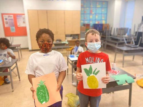 Two young children holding up pieces of art they made in a classroom.