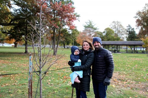 Two parents and their baby stand next to a newly planted tree.