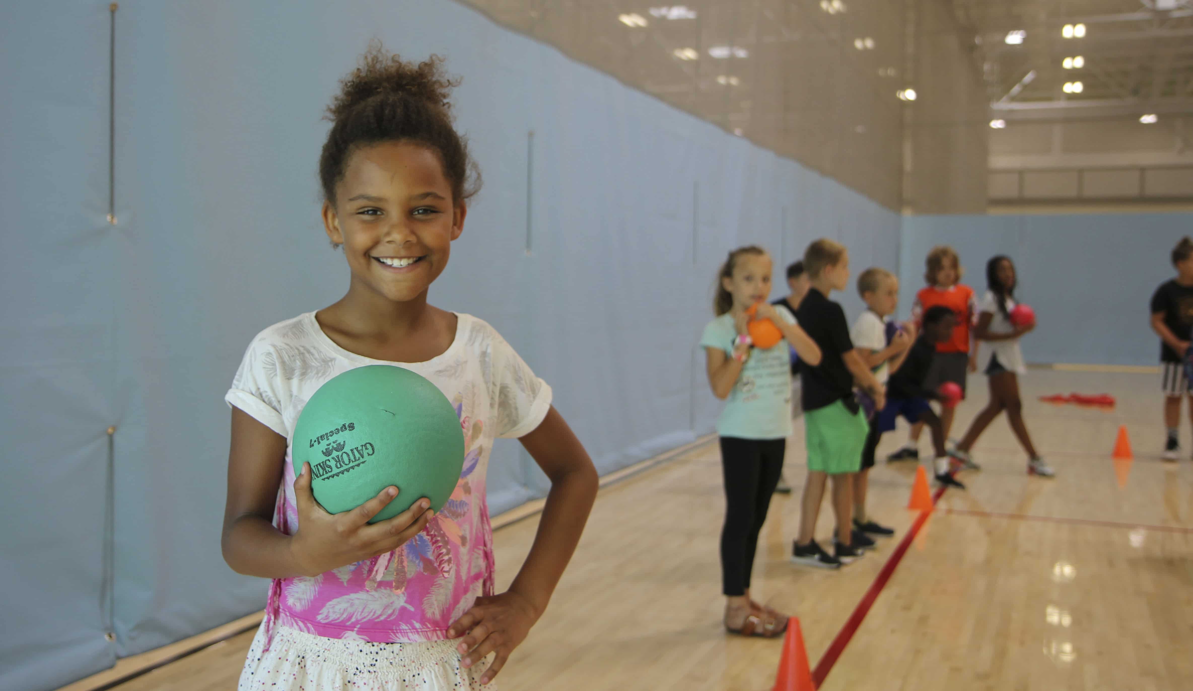 A child is holding a ball and smiling at the camera. There is a line of children behind her.