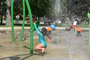 Child hanging from outdoor Splash Pad equipment being sprayed with water for fun.