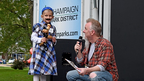 Little girl wearing flannel dress and fake black mustache holding a balloon animal.