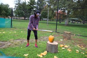 Person wearing flannel swinging bat at a pumpkin to smash it with a smile on their face.