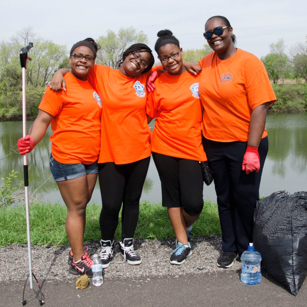 4 women in matching orange shirts stand in front of Kaufman Lake during Boneyard Creek Community Day.