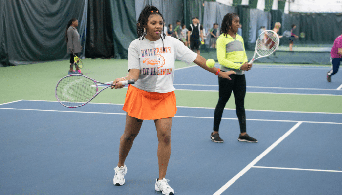 Two women stand on a tennis court, racquets in hand. A tennis ball flies toward the first woman's racquet.
