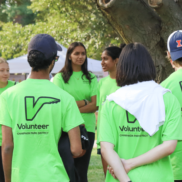 A group of young adults stand in a circle. They are wearing matching green shirts with the word "Volunteer" printed on the back.
