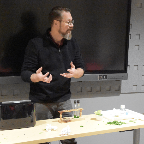 A person stands in front of a classroom with science experiments on the table in front of him.